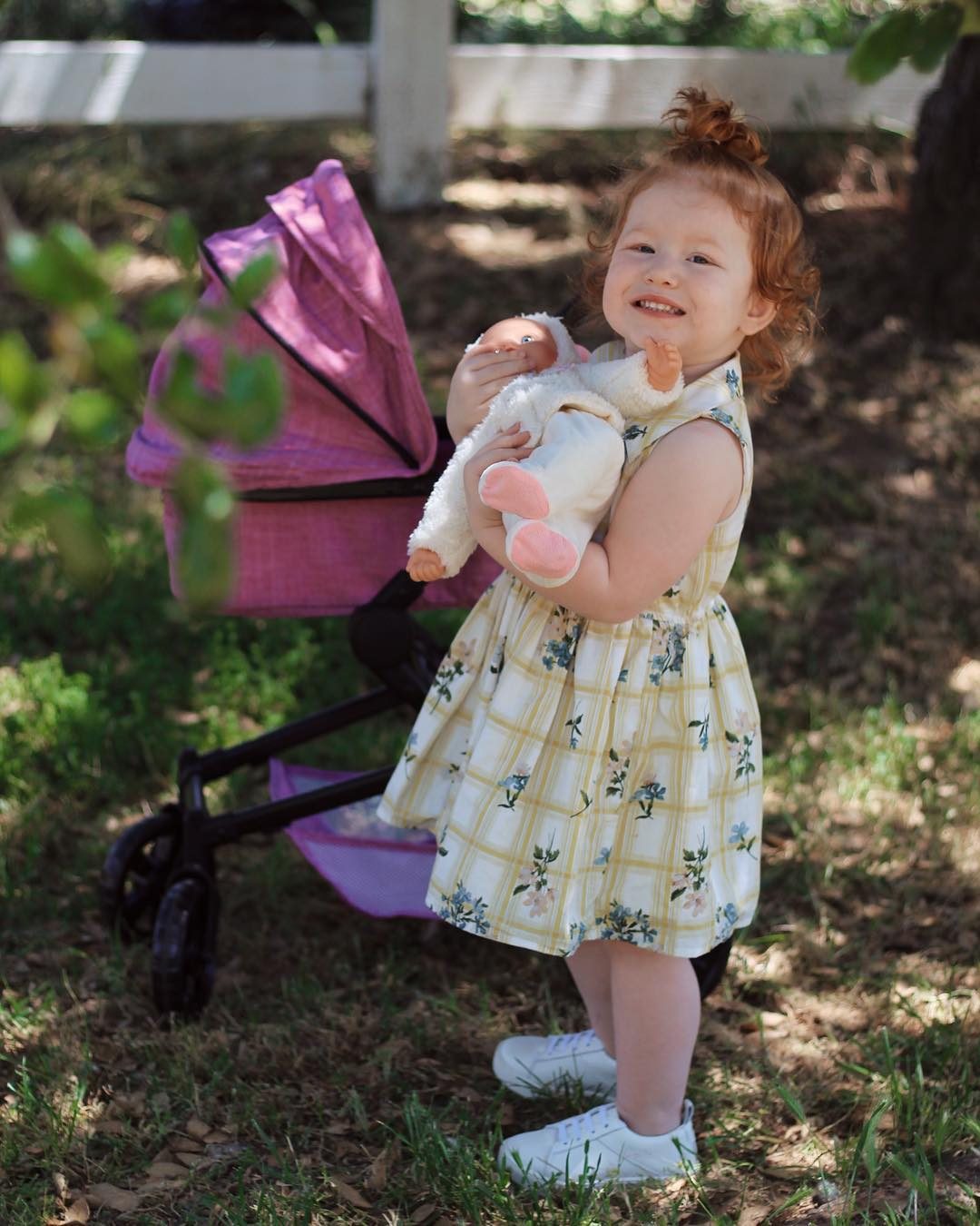 A toddler in florals holds a soft doll beside her purple pram